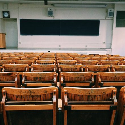 Several rows of wooden chairs face a podium and blackboard at the front of an empty classroom.
