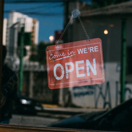 A sign that reads, "Come In We're Open" is hanging on the inside of a glass door and is being viewed from outside.