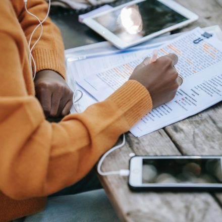 A black woman in an orange sweater is highlighting text on a printed piece of paper. To her right is a smartphone with a cord for headphone going towards her head. On her left is a tablet computer.