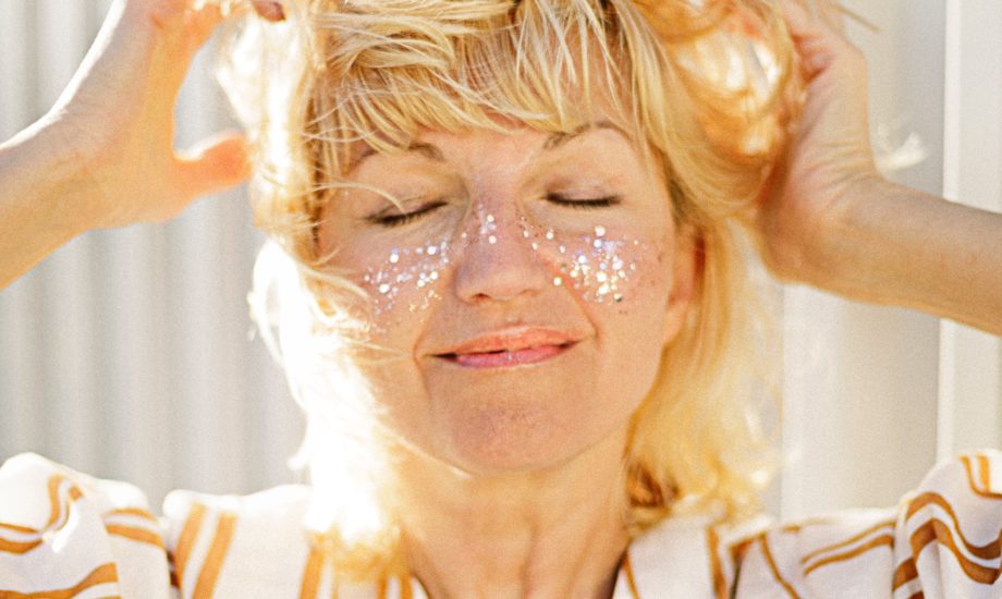 A blonde woman wearing a white shirt with orange stripes has glitter on her cheeks and is smiling.