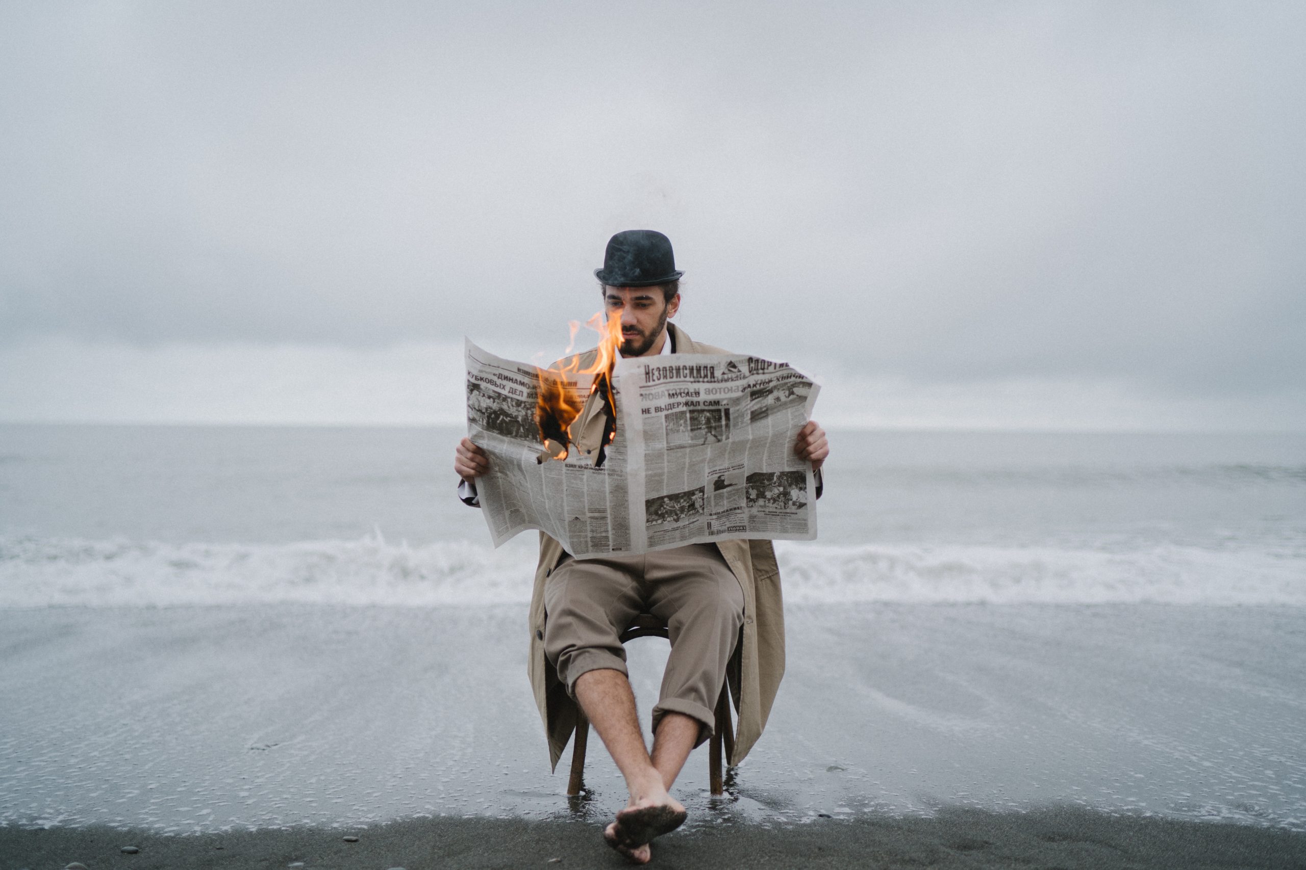 A photo of a man in a tan trenchcoat and a black bowler hat sits on a chair on a beach, and is holding a newspaper open and it turns to flame.
