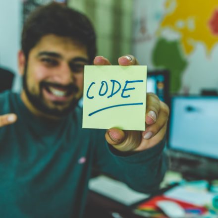 A bearded man in a grey sweater is holding a yellow sticky note which has CODE written upon it.