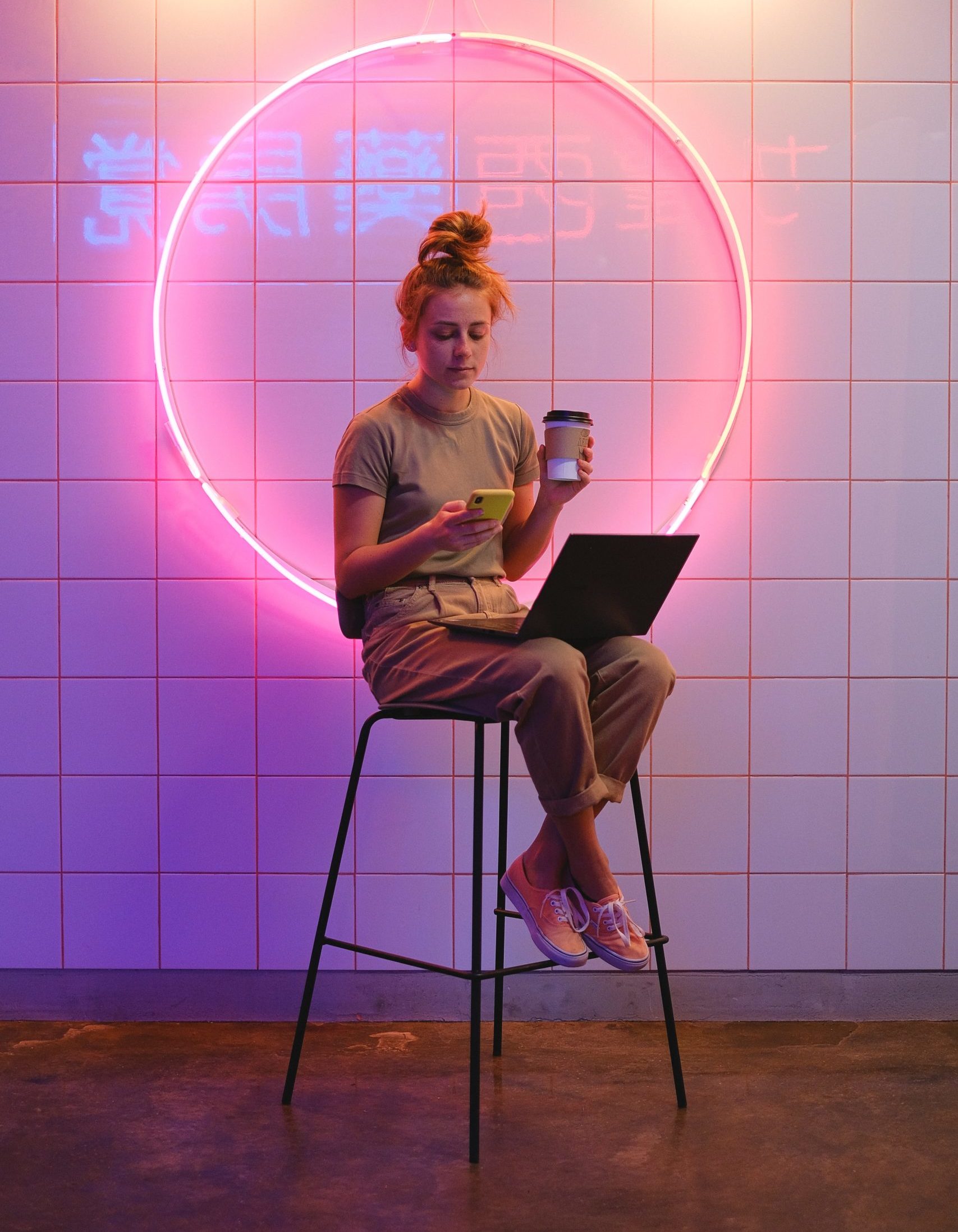 A photo of a woman sitting on a stool in front of a white tiled wall. A large neon circle is hanging on the wall and neon chinese characters are reflected in the glaze of the tiles. The woman is holding a coffee cup in one hand, a smartphone in the other, and has a laptop on her lap.