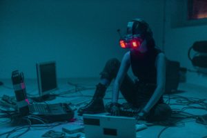 A color photo os a person wearing VR goggles while sitting on the floor in a darkened room. They are surrounded by several cables and various computers.