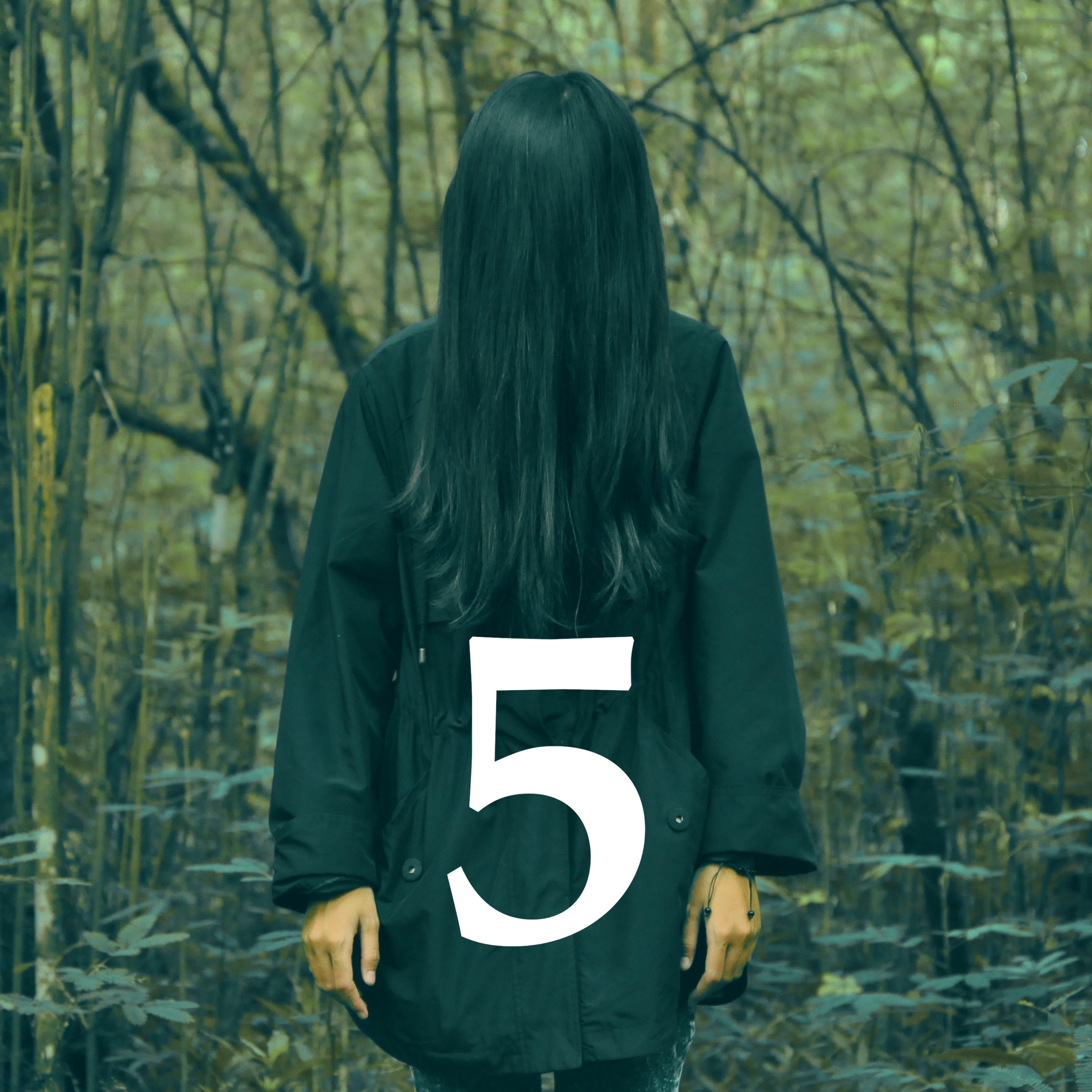 A color photo of a person in a black coat standing in a forest as their long black hair obscures their face.