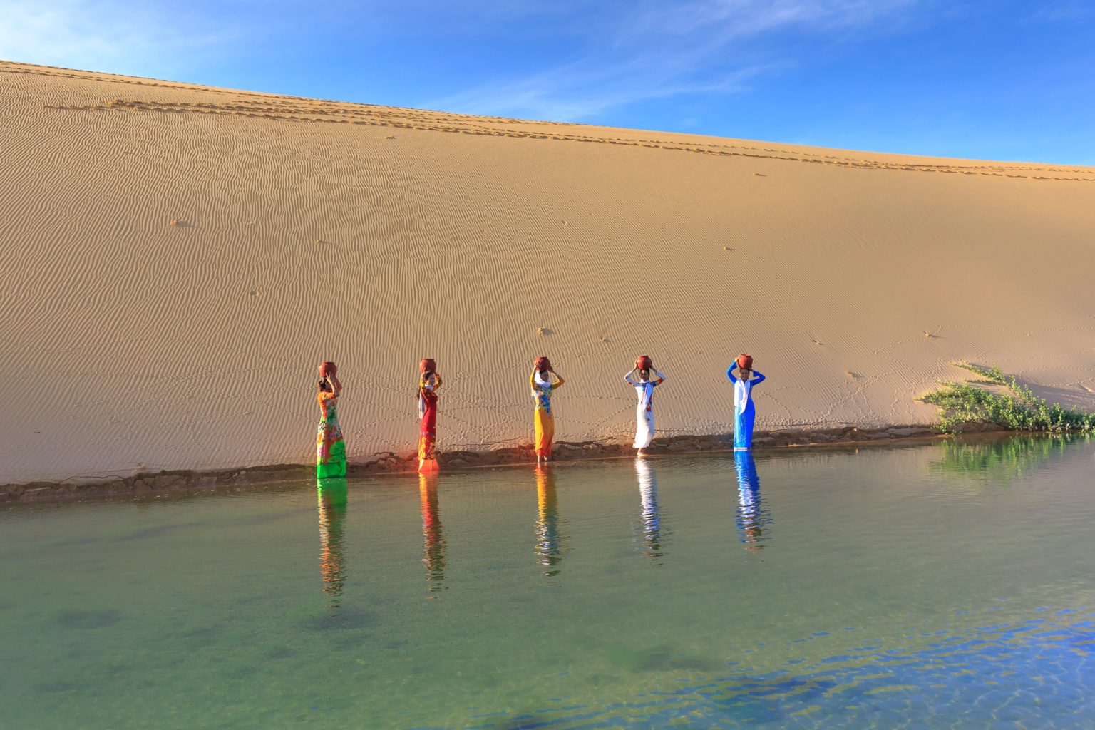 A color photo of five women in brightly colored dresses carrying jugs of water on their heads. A sand dune is in the background while an oasis of water is in the foreground, with the women standing at the edge of the water.