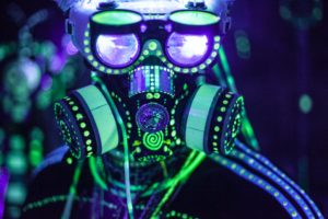 A person wearing a gas mask and eye goggles festooned with fluorescent green dots that are illuminated under a blacklight.